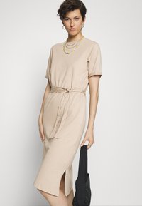 Vestido bege de comprimento médio feito de algodão suave, com mangas curtas dobradas, cintura com cinto e uma fenda de um lado.
