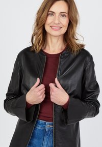 Musta nahkatakki ilman kaulusta, vetoketjukiinnitys ja löysät hihat, päällä marooni paita. Malli hymyilee, kädet takin lähellä.