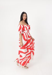 Maxikleid mit schulterfreiem Schnitt, roten und weißen abstrakten Mustern, gesmokt Oberteil und fließendem Rock. Getragen mit goldenen offenen Zehenabsätzen.