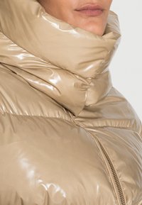 Kurtka puchowa w jasnym odcieniu beżu, z wysokim połyskiem i teksturowanym wykończeniem. Kołnierz jest duży i złożony, z detalem w postaci suwaka.