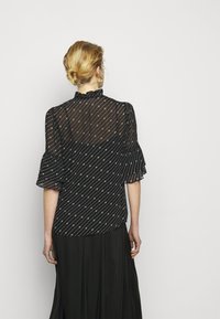 Blusa nera in chiffon con collo arricciato, maniche corte e motivi bianchi diagonali. La vista posteriore mostra un design fluido abbinato a una gonna nera.