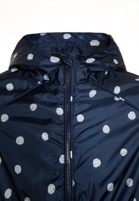 Marinblå vattentät jacka med dragkedja, prydd med vita prickar och en huva. Tillverkad av mjukt, lätt material.