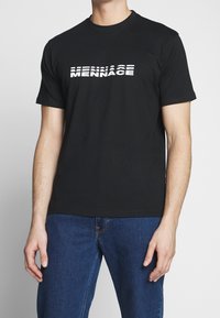 T-shirt en coton noir avec un imprimé textuel blanc disant "MENNACE" avec des épaisseurs variées, encolure ronde, manches courtes et coupe décontractée.