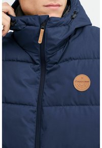 Blaue gepolsterte Jacke mit hohem Kragen und Reißverschluss. Verfügt über ein kreisförmiges, hellbraunes Logo-Patch und strukturierter Stoff für einen eleganten Look.