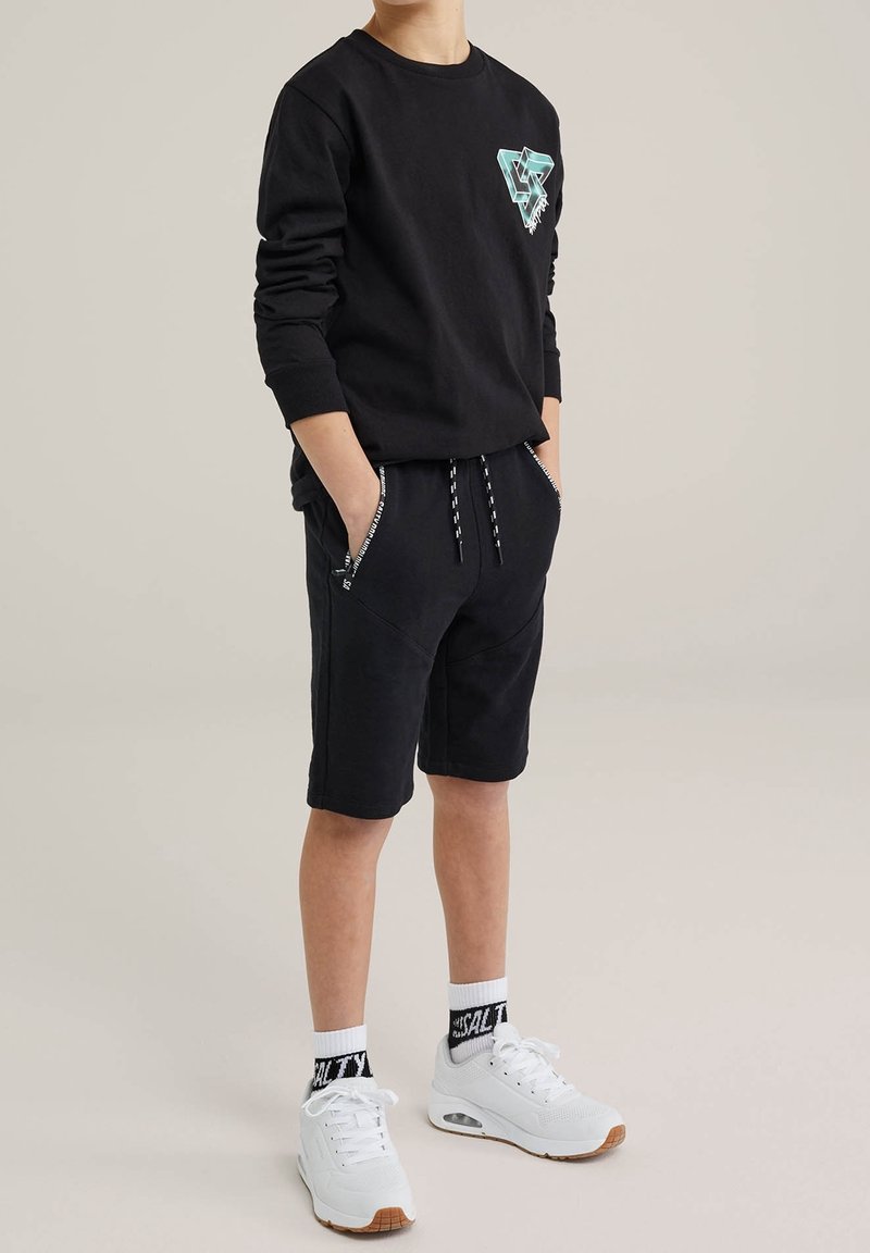 Enfant portant un t-shirt noir à manches longues avec un motif, un short noir avec un cordon, des baskets blanches et des chaussettes blanches, debout les mains dans les poches.
