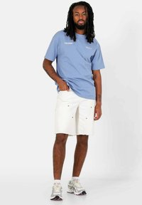 Lichtblauw katoenen T-shirt met witte logotext, gecombineerd met witte denimshorts met zakdetails en een rafelige zoom. Grijze en roze sneakers.