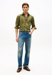 Πουκάμισο σε ελιά πράσινο με κουμπιά, ανοιχτό μπλε τζιν με ίσια γραμμή, καφέ παπούτσια. Το σύνολο διαθέτει ορατές ραφές και διακριτή υφή υφάσματος.