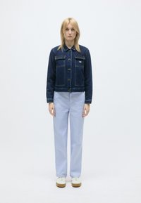 Tamna plava traper jakna s prednjim džepovima, kopčanjem na dugmad i žutim šavovima; uparena s svijetloplavim hlačama i bijelim tenisicama.