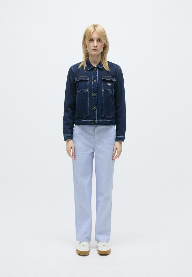 Donkerblauwe spijkerjack met voorzakken, knoopsluiting en gele stiksels; gecombineerd met lichtblauwe broek en witte sneakers.