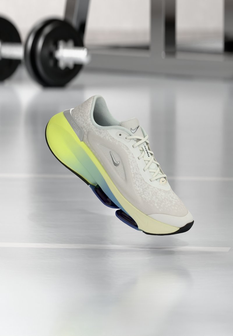Chaussure de course Nike gris clair et blanche avec une semelle amortissante jaune et bleue, flottant au-dessus d’un sol de gym réfléchissant avec du matériel de musculation en arrière-plan.