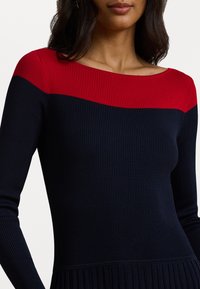 Vestido canelado com corpo azul-marinho e painel de ombro vermelho. Apresenta um decote barco e mangas compridas. Textura plissada na bainha.