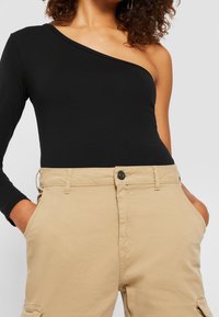 Musta yksiolkaiminen bodysuit, joka on yhdistetty vaaleanbeigeihin korkeavyötäröisiin cargo-housuihin, joissa on sivutaskut ja napitus. Sileä pehmeä rakenne ja istuva muotoilu.