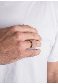 Inel de aur cu un model geometric purtat pe o mână, având un finisaj neted. Mâna se odihnește pe un tricou alb.