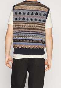 Pletená vesta s vícebarevnými geometrickými vzory v hnědé, modré a šedé barvě s námořnicky modrým lemováním; nošená přes světlou košili.