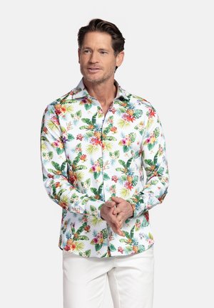 Man draagt een wit shirt met lange mouwen met kleurrijke tropische bloemen- en ananasprint, gecombineerd met een witte broek, staand tegen een effen achtergrond.