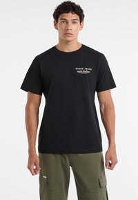 Młody mężczyzna z kręconymi włosami, noszący czarną koszulkę Guess Jeans i oliwkowe spodnie cargo, stoi na tle białym.