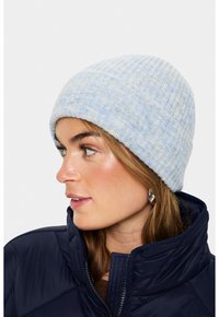 Žena v světle modré pletené čepici a námořnickém bundě, dívající se vlevo, se stříbrnými kruhovými náušnicemi viditelnými.