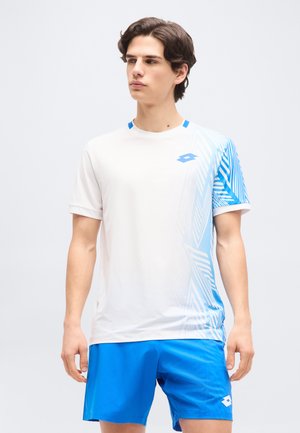 Νέος άντρας που φοράει λευκό και μπλε αθλητικό πουκάμισο με γεωμετρικό μοτίβο και αντίστοιχα μπλε σορτς, όρθιος μπροστά σε απλό φόντο.