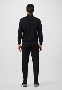 Muškarac stoji okrenut leđima, nosi crnu sportsku jaknu i hlače s logotipom na ovratniku jakne, protiv jednostavne svijetle pozadine.