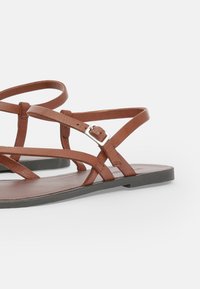 Sandali piatti in pelle marrone con cinturini incrociati, fibbia regolabile e design minimalista della suola piatta.