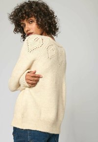 Pull crème en maille texturée, avec un motif de feuilles en dentelle au dos, et des poignets et un ourlet côtelés. Design ajusté.