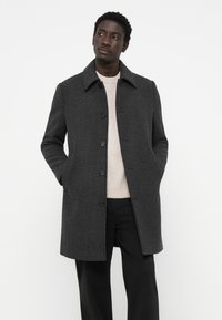 Férfi sötétszürke kockás zakót visel világos bézs pulóver és fekete nadrág felett, kezei a zsebében, fehér háttér előtt áll.