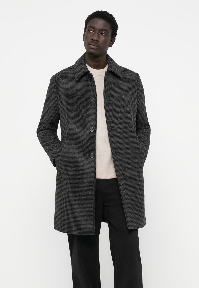 Homme portant un manteau à carreaux gris foncé boutonné sur un pull beige clair et un pantalon noir, se tenant les mains dans les poches sur un fond blanc.