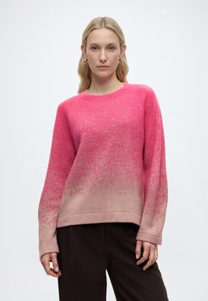 Nő, aki hosszú ujjú, rózsaszín átmenetes pulóvert és sötét nadrágot visel, semleges arckifejezéssel áll egy egyszerű háttér előtt.