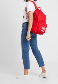 Red backpack made of durable material, featuring a front zip pocket, adjustable straps, and white Adidas logo. Accompanied by blue jeans and white sneakers.