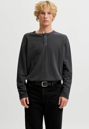 Jeune personne aux cheveux blonds bouclés portant un t-shirt henley à manches longues gris foncé et un pantalon noir avec ceinture, debout devant un fond uni.