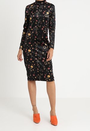 Schwarzes geblümtes Midikleid mit langen Ärmeln, getragen mit leuchtend orangefarbenen Slip-On-Schuhen, gezeigt an einer stehenden Person vor einfachem weißem Hintergrund.