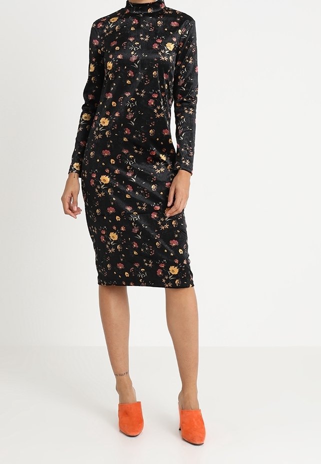 Robe midi noire à fleurs à manches longues, portée avec des chaussures slip-on orange vif, présentée sur une personne debout sur fond blanc uni.