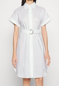 Vestito bianco a camicia con maniche corte, apertura frontale con bottoni, vita regolabile con una cintura dotata di fibbia argentata e una consistenza di cotone liscia.