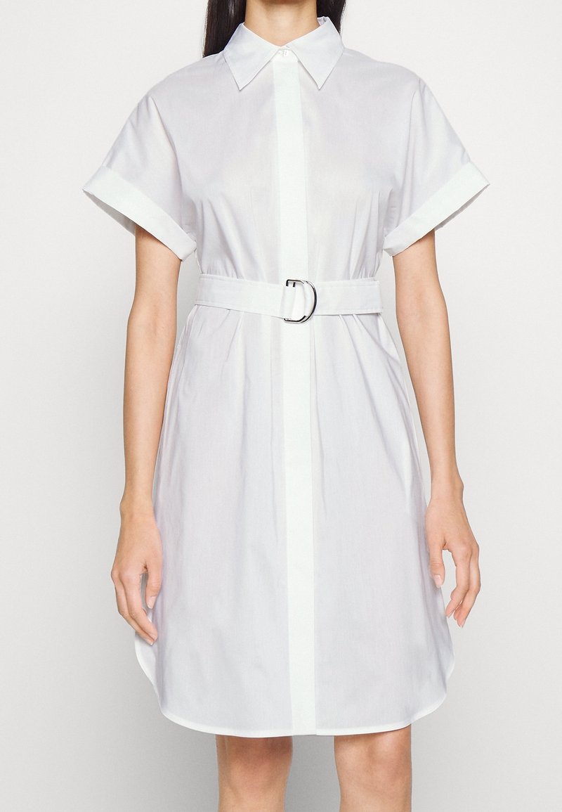 Vestito bianco a camicia con maniche corte, apertura frontale con bottoni, vita regolabile con una cintura dotata di fibbia argentata e una consistenza di cotone liscia.