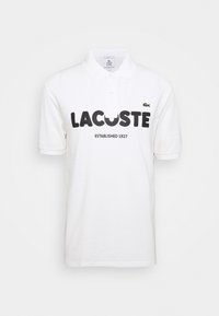 Polo shirt blanc à manches courtes avec le texte noir "LACOSTE", petit logo crocodile vert sur la poitrine, et "ÉTABLI EN 1927" en dessous du texte.