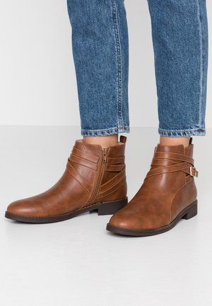 Bottines en cuir marron avec fermeture éclair latérale et sangles décoratives portées avec un jean bleu sur un fond uni clair.