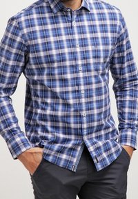 Mann trägt ein blau-weiß kariertes Hemd mit langen Ärmeln, die Hände in den Taschen einer dunklen Hose, zugeschnitten auf Kinn- und Oberschenkelhöhe.