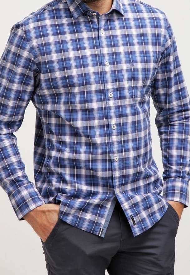 Mann trägt ein blau-weiß kariertes Hemd mit langen Ärmeln, die Hände in den Taschen einer dunklen Hose, zugeschnitten auf Kinn- und Oberschenkelhöhe.