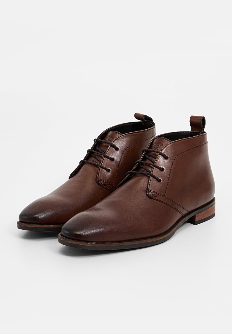 Botas de tobillo de cuero marrón con un diseño elegante, puntera redonda, cordones en el frente y un pequeño tacón. Incluyen una pestaña de agarre en la parte posterior para facilitar el calzado.