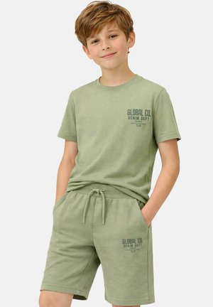 Jongen met kort lichtbruin haar die een bijpassend lichtgroen T-shirt en korte broek draagt met de tekst "GLOBAL CO. DENIM DEPT" op beide.