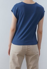 Person som bär en blå kortärmad skjorta och beige byxor, står vänd bort mot en helt vit vägg som bakgrund.