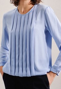 Blusa azzurro chiaro a maniche lunghe con design frontale a pieghe. Tessuto liscio, scollatura rotonda e polsini abbottonati. Fit su misura con texture morbida.