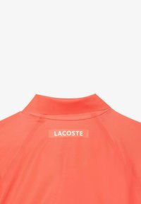 Orange polo skjorta med krage. Har en vit "LACOSTE"-logotyp på en rektangulär etikett på baksidan. Slät tygtextur.
