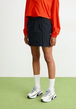 Osoba nosząca czarną spódnicę Nike, białe tenisówki, białe wysokie skarpety i pomarańczowy top z długim rękawem, stojąca na zielonej podłodze na tle białej ściany.
