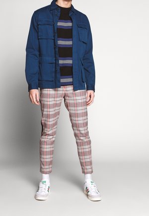 Mann trägt blaue Jacke, schwarz-blau gestreiftes Hemd, beige karierte Hose, weiße Socken und weiße Turnschuhe mit grünen und roten Streifen.