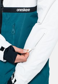 Türkis-weiße Skijacke mit verstellbaren Manschetten; verfügt über eine Reißverschlusstasche mit weißem Zugband und kontrastierendem schwarzem Logo auf der Brust.