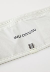 Biely textúrovaný materiál s logom "SALOMON" v sivej farbe a malou čiernou a sivou textilnou slučkou pri okraji.