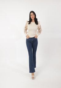 Maglione beige chiaro a coste con maniche in pizzo, abbinato a jeans a zampa blu. Modella che indossa sandali con tacco, in posa con le mani nelle tasche.