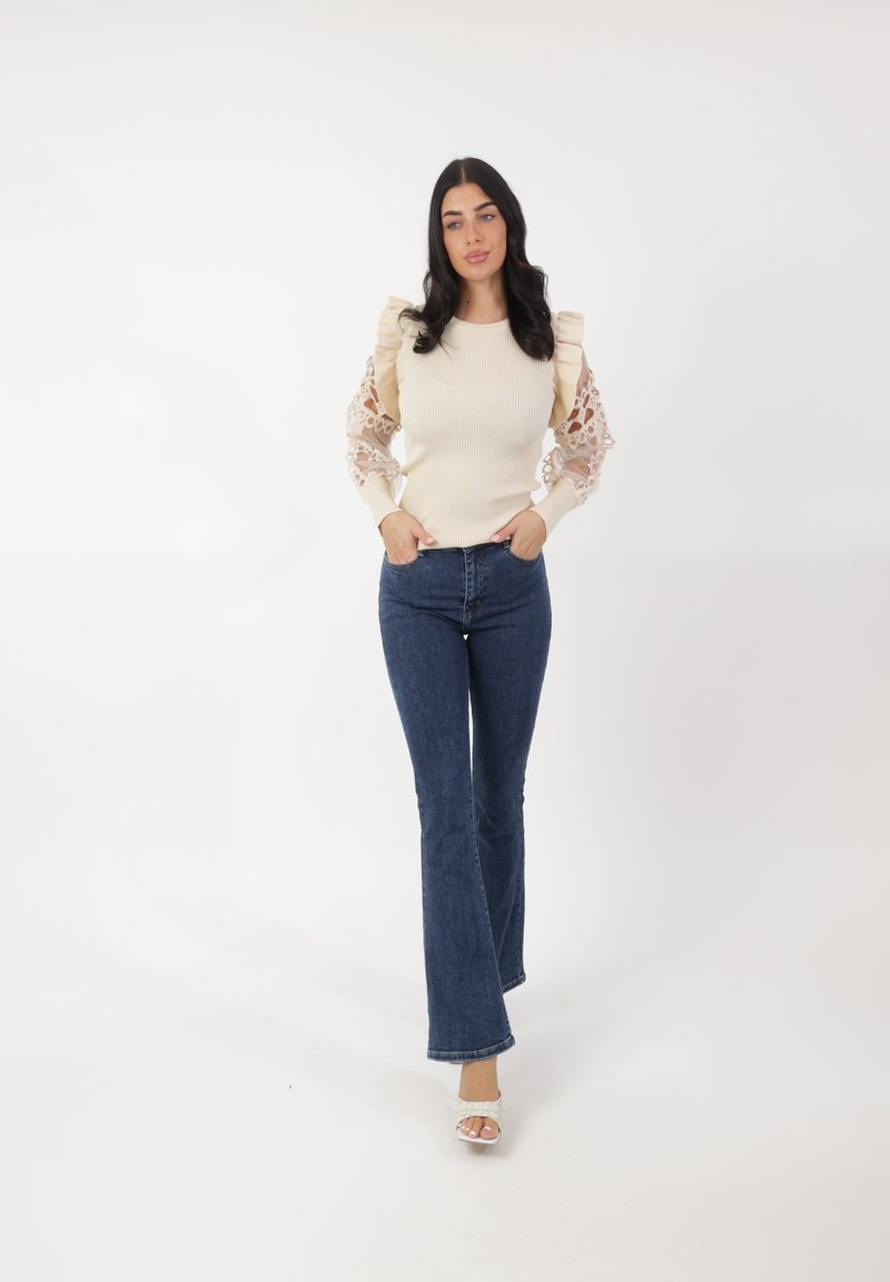 Maglione beige chiaro a coste con maniche in pizzo, abbinato a jeans a zampa blu. Modella che indossa sandali con tacco, in posa con le mani nelle tasche.
