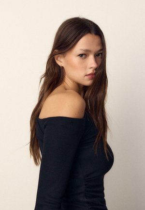 Mlada žena s dugom smeđom kosom koja nosi crnu majicu s otkrivenim ramenima, gleda preko ramena na jednobojnoj pozadini.